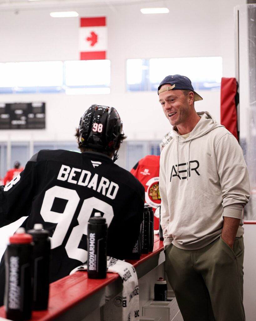 Jonathan Toews talks with Connor Bedard on the bench at Blackhawks Ice Center before the home opener